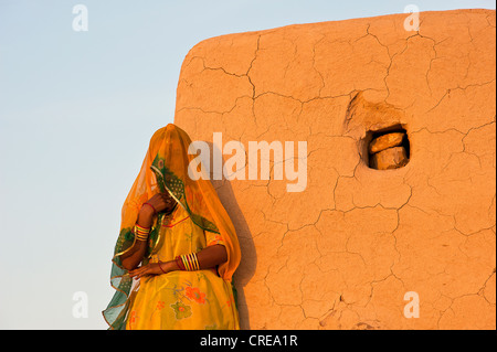 Eine verschleierte junge indische Frau trägt einen gelben Sari steht vor einer Wand in der Sonne, Thar-Wüste, Rajasthan, Indien, Asien Stockfoto