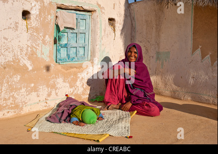 Eine freundliche indische Frau trägt einen roten Sari sitzen auf dem Boden in ihrem Innenhof, liegt ihr schlafendes Kind vor ihr auf Stockfoto