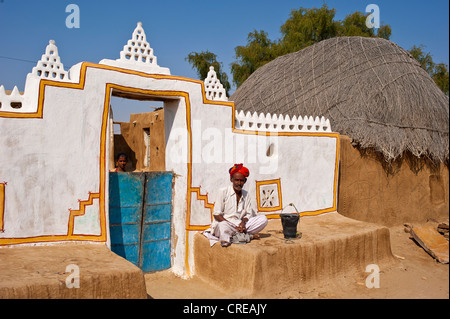 Rajasthani, indischer Mann trägt einen Dhoti und Turban, sitzt vor seinem traditionell gebaut und bemalt Haustür Stockfoto