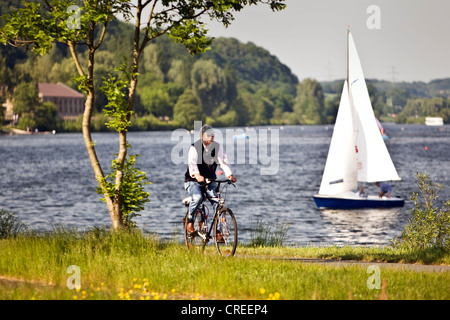 Mann auf einem Fahrrad an den Kemnader Stausee mit Segelboot, Witten, Ruhrgebiet, Nordrhein-Westfalen, Deutschland Stockfoto