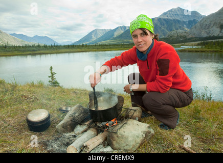 Junge Frau auf einem Lagerfeuer kochen in einem dampfenden Topf, camping, Mackenzie Mountains hinter, Wind River, Peel Watershed Lenkung Stockfoto