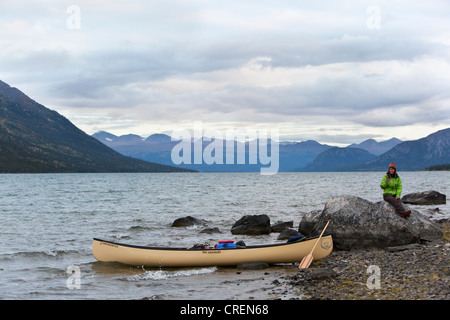 Junge Frau sitzt auf einem Felsen, Kanu und hölzernen Paddel auf dem Ufer, Kusawa See, Bergen im Hintergrund, Yukon Territorium, Kanada Stockfoto