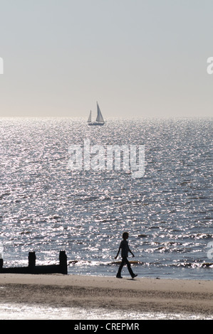 Eine Frau geht am Rand des Meeres an einem Strand in den frühen Morgenstunden, mit einem Segelboot in der Ferne Frinton Essex Stockfoto