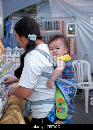 Schlinge für den Kinderrücken. Mutter im thailändischen Marktstallhalter, der Baby auf dem Rücken trägt. Thailand S.E. Asien Stockfoto