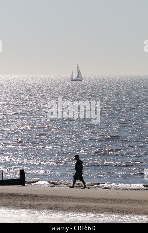 Ein Mann geht am Rand des Meeres an einem Strand in den frühen Morgenstunden, mit einem Segelboot in der Ferne Frinton Essex Stockfoto