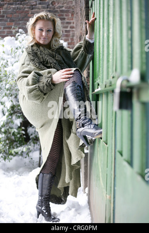 blonde Frau trägt grüne Wollmantel und schwarze Stiefel am Tor Stockfoto