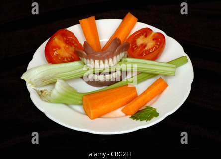 Ein Stück Sellerie zwischen die Zähne aus einer Reihe von Zahnersatz, auf einen Teller voll mit verschiedenen anderen Gemüsesorten. Stockfoto