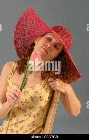 junge schöne rothaarige Frau mit großen roten Hut, riecht um eine Tulpe Stockfoto