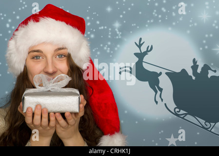 Junge Frau in Nikolausmütze Weihnachtsgeschenk, Grafik der Schlitten des Weihnachtsmannes im Hintergrund halten Stockfoto