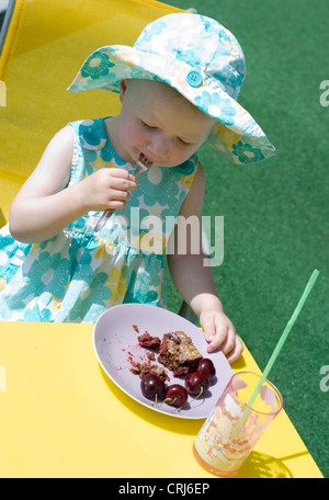 kleines Mädchen im Sommer Tuch mit Sonnenhut sitzen am Tisch für Kinder im Garten Kirsch Kuchen essen Stockfoto