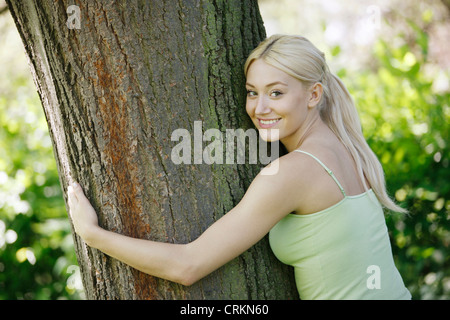 Eine junge, blonde Frau umarmt einen Baum lächelnd Stockfoto
