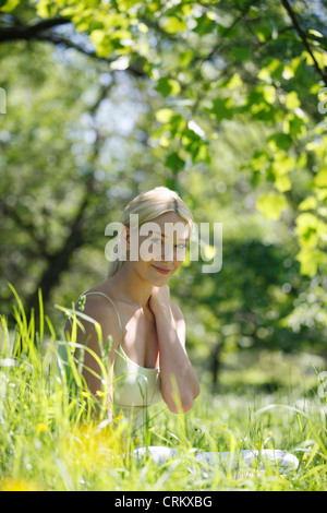 Eine junge, blonde Frau sitzt in der Wiese, Lächeln und Kamera Stockfoto