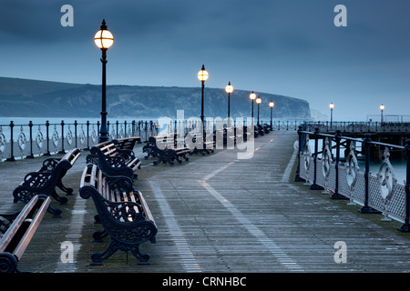 Bänken unter Lampen entlang Swanage Pier in der Dämmerung. Stockfoto