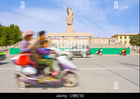 Mit Bewegungsunschärfe.  Fahrzeuge und Personen reisen Sie vorbei an der Statue von Mao Zedong gegenüber dem Stadtplatz in Kashgar. Stockfoto