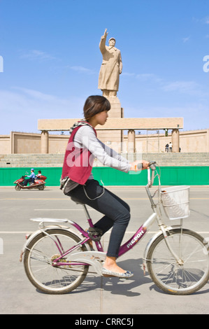 Mit Bewegungsunschärfe.  Eine junge Chinesin auf auf einem Fahrrad fahren Sie vorbei an der Statue von Mao Zedong in Kashgar. Stockfoto