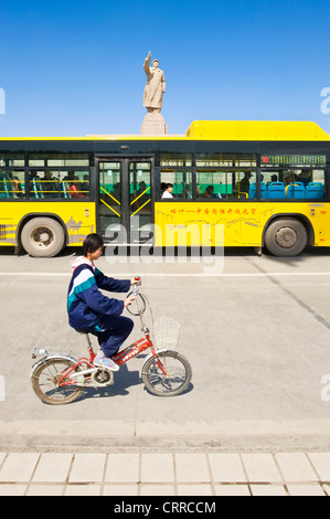 Mit Bewegungsunschärfe.  Eine junge chinesische Schulmädchen auf auf einem Fahrrad fährt vorbei an der Statue von Mao Zedong in Kashgar. Stockfoto