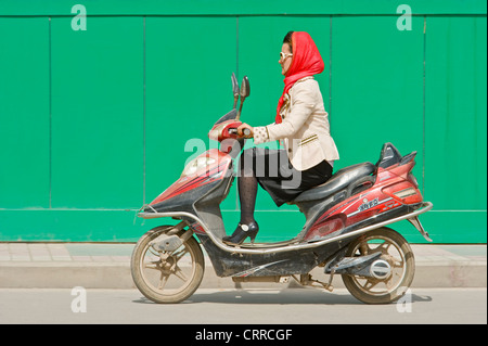 Eine chinesische Frau reist entlang einer Straße auf ihrem Roller in Kashgar. Stockfoto