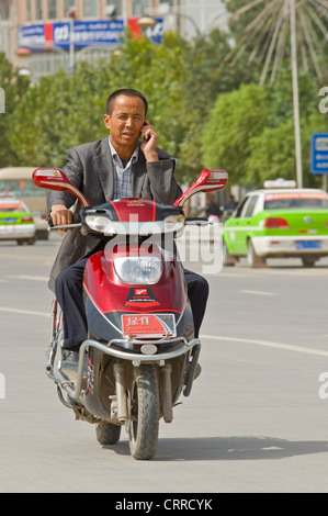 Ein chinesischer Mann reist entlang einer Straße auf seinem Roller während des Gesprächs auf seinem Handy in Kashgar. Stockfoto