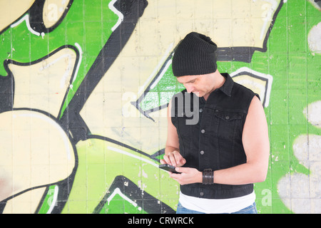 Mann trägt eine Mütze und stand vor Graffiti-Wand sendet eine Nachricht auf seinem Handy Stockfoto