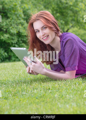 Glückliche junge weibliche Erwachsene mit roten Haaren liegen auf einer Wiese mit einem TabletPC Stockfoto