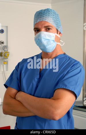 Ein männlicher Arzt in blauen OP-Kittel, Haube und Maske, steht mit verschränkten Armen. Stockfoto