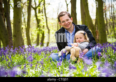 Vater und Tochter sitzen in einem Park, umgeben von Glockenblumen, hält ein Mädchen einen Teddybär. Stockfoto