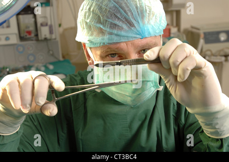 Ein maskierter Chirurg hält ein paar Pinzette und Skalpell in seinen behandschuhten Händen. Stockfoto