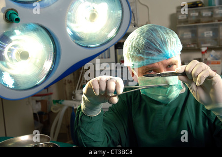 Ein Chirurg in vollständige chirurgische Kleid hält eine Scaplel und ein paar Zangen in einem Operationssaal. Stockfoto