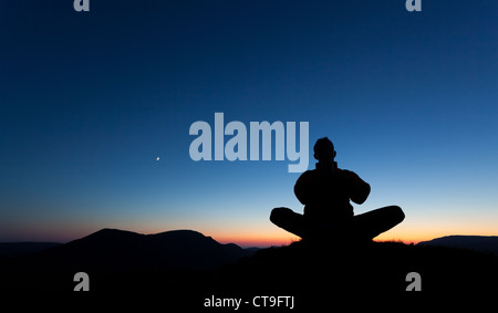 Mann, der auf dem Gipfel eines Berges bei Sonnenuntergang mit dem Mond am Himmel betet. Stockfoto