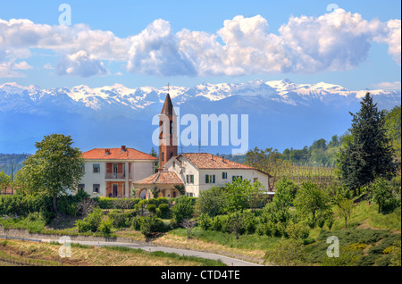 Landhäuser und Kirchlein unter grünen Bäumen und blauen Berge mit schneebedeckten Gipfeln im Hintergrund im Piemont, Norditalien. Stockfoto