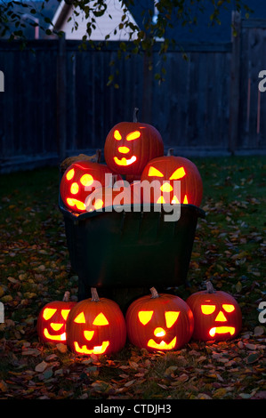 Geschnitzte Kürbisse beleuchtet in einer Schubkarre mit Herbstlaub auf Hinterhof Rasen in der Dämmerung. Stockfoto