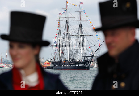 United States Coast Guard, die Seeleute in 1812 Uniformen auf einem Pier stehen, als die USS Constitution, Hintergrund Mitte, im Hafen von Boston Segel Stockfoto