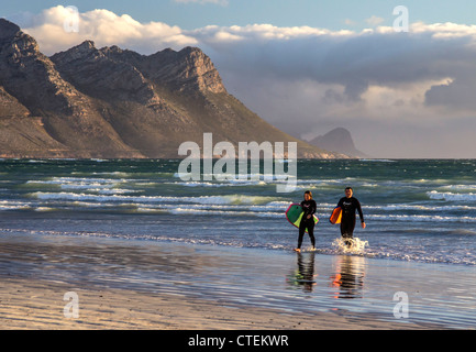 Zwei Surfer in nassen Anzügen, die Surfbretter tragen, tauchen aus dem Meer auf - Das Strandbad Strand, Westkappo, Südafrika Stockfoto