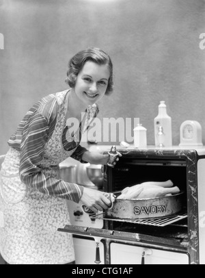 1930ER JAHRE HAUSFRAU IN SCHÜRZE AUFNAHME DER TÜRKEI IN PAN LESEN BOHNENKRAUT AUS OFEN BLICK IN DIE KAMERA Stockfoto