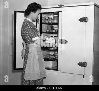 1930ER JAHRE HAUSFRAU SCHÜRZE NEHMEN EIER AUS SCHÜSSEL VON ICEBOX Stockfoto