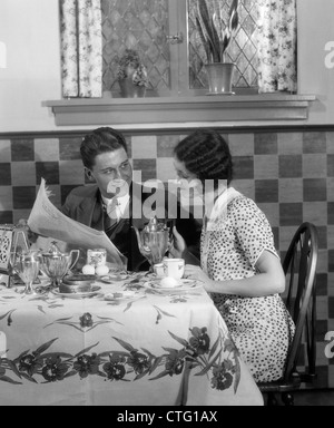 1920S 1930S ZU ZWEIT AM FRÜHSTÜCKSTISCH MIT BLICK AUF ZEITUNG FRAU IMMER BEREIT ZUM GIEßEN KAFFEE AUS SILBERNEN TOPF Stockfoto