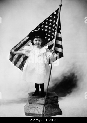 1890ER JAHREN JAHRHUNDERTWENDE LÄCHELNDE MÄDCHEN STEHEN AUF PLATTFORM TRÄGT EIN WEIßES KLEID UND EINEN DUNKLEN HUT HÄLT EINE AMERIKANISCHE FAHNE Stockfoto