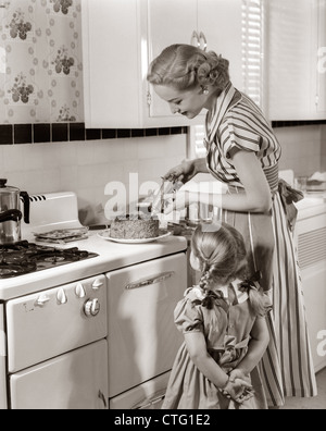 1950ER JAHRE HAUSFRAU IN DER KÜCHE DEKORATION KUCHEN AUF HERD MIT GEBÄCK GUN WÄHREND TÖCHTERCHEN UHREN Stockfoto