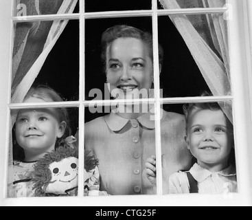 1950ER JAHREN MUTTER MIT ZWEI KINDERN SUCHEN FENSTER IN RICHTUNG BETRACHTER Stockfoto