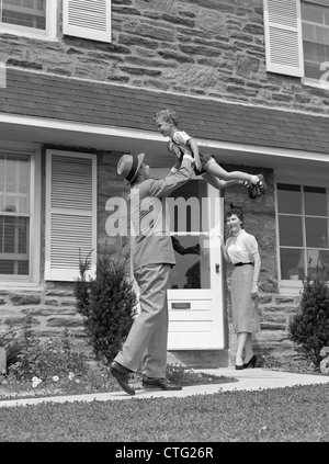 1950S 1960S VATER HEIMKEHREN HEBEN SOHN IN LUFT WÄHREND FRAU AUS TÜR SCHAUT Stockfoto