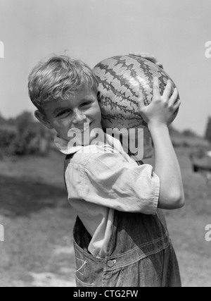 1930ER JAHREN BAUERNJUNGE IN OVERALLS TRAGEN WASSERMELONE AUF SCHULTER Stockfoto