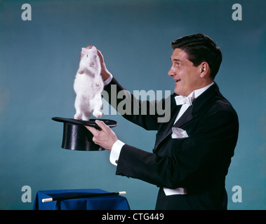 1960ER JAHREN MAGIER TRÄGT SCHWARZEN SMOKING LÄCHELND ZIEHENDE WEIßE KANINCHEN AUS ZYLINDERHUT Stockfoto