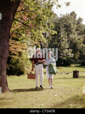 1960ER JAHREN LÄCHELND ÄLTERES PAAR GEHEN ARM IN ARM MIT PICKNICK-KORB Stockfoto