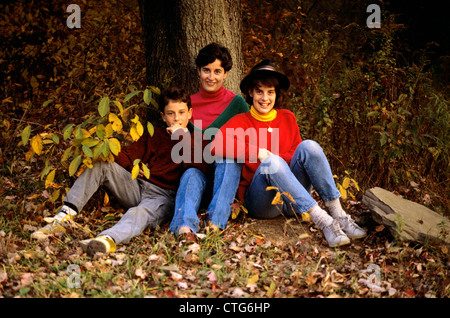 1980ER JAHRE FAMILIE MUTTER SOHN UND TOCHTER SITZEN AUF BASIS DER BAUM IM WALD HERBST Stockfoto