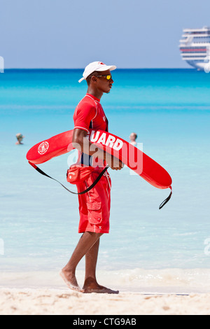 Junge schwarze männliche Rettungsschwimmer am Strand zu Fuß mit Schwimmer Rettungsgerät im Half Moon Cay, Bahamas Stockfoto