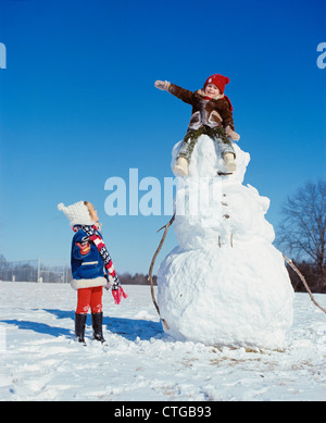 1970ER JAHRE BOY SITTING ON TOP OF SCHNEEMANN KOPF WINKEN FÜR MÄDCHEN Stockfoto