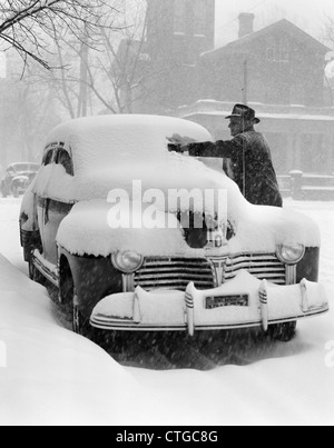 1940ER JAHRE MANN ENTFERNEN SCHNEE VON WINDSCHUTZSCHEIBE PARKENDEN AUTOS Stockfoto