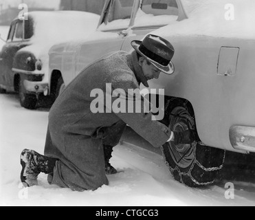 1950ER JAHRE MANN IN MANTEL & HUT KNIETE IM SCHNEE ANPASSEN AUF REIFEN-KETTEN Stockfoto