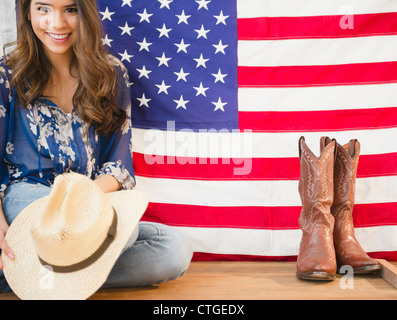 Hispanic Frau Cowboyhut halten und sitzen neben der amerikanischen Flagge Stockfoto