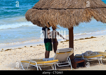 Paar am Strand entlang der Riviera Maya, Yucatan, Mexiko. Karibischen Meer steht hinter ihnen, wie sie unter einer Palme Stroh Schutz stehen. Stockfoto